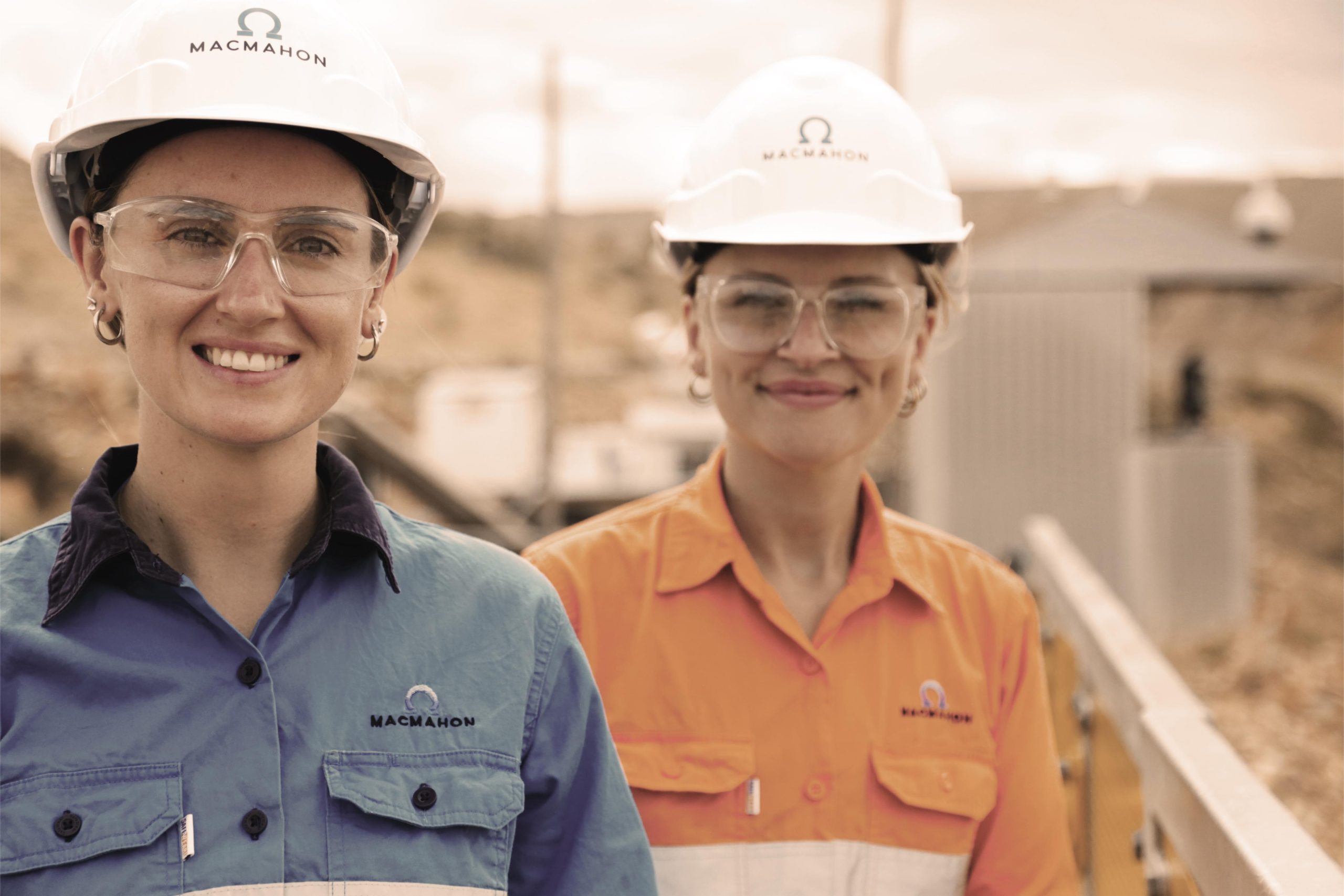 Two smiling female Macmahon workers in hard hats outdoors