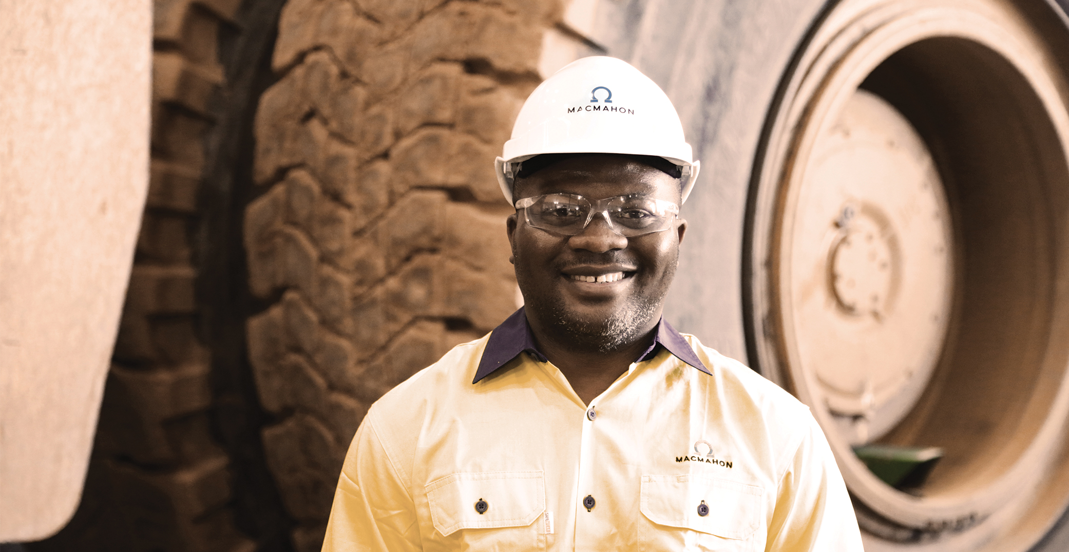 Smiling Macmahon worker in hard hat next to large truck tires