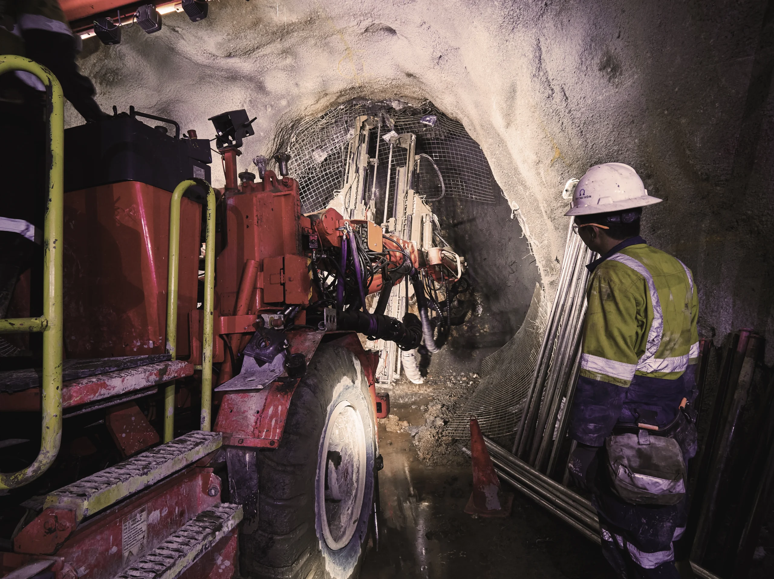 Worker observing red drill jumbo in underground tunnel